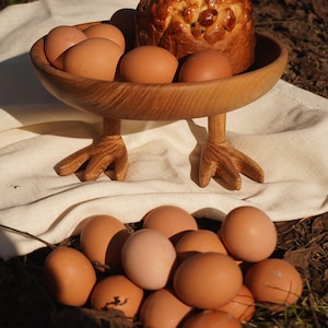 May include: A wooden bowl with chicken feet legs, filled with brown eggs and a loaf of bread. The bowl sits on a white cloth, with more eggs scattered below. The wood has a natural grain.