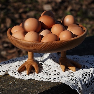 May include: A wooden bowl filled with brown eggs, set on a white lace doily. The bowl has three-toed wooden feet, giving it a rustic look. The eggs are light brown. The bowl is likely made of wood.