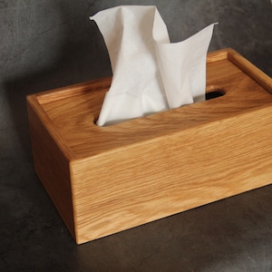 May include: A wooden tissue box holder with a natural wood finish. The box has a slot for pulling out tissues and is made of solid wood.