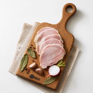 May include: A wooden serving board with a handle holds four thin slices of pink ham, two cloves of garlic, a halved radish, and several dried bay leaves. The board rests on a piece of printed newspaper, featuring a small circular logo at the bottom.