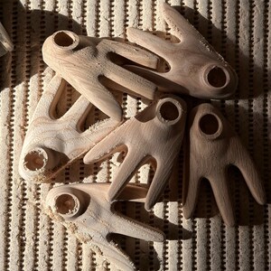 May include: A collection of unfinished wooden craft pieces, each with a unique shape featuring a central hole and three finger-like projections. The natural wood grain is visible, and the pieces are scattered on a textured surface.