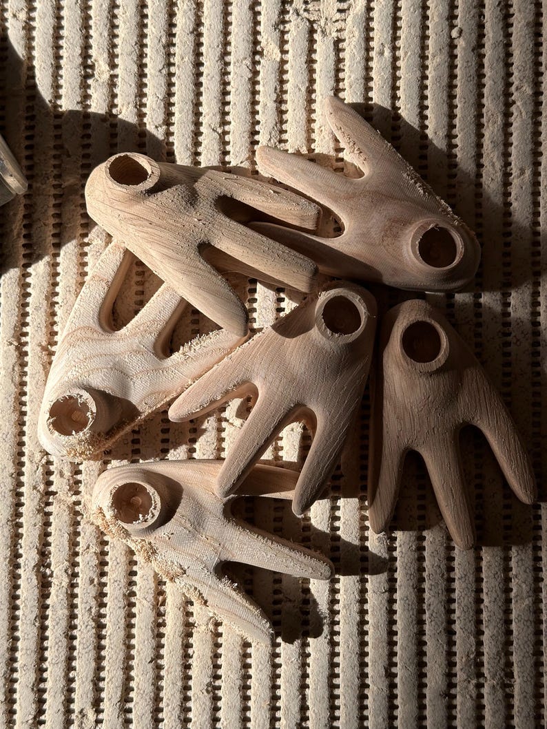 May include: A collection of unfinished wooden craft pieces, each with a unique shape featuring a central hole and three finger-like projections. The natural wood grain is visible, and the pieces are scattered on a textured surface.