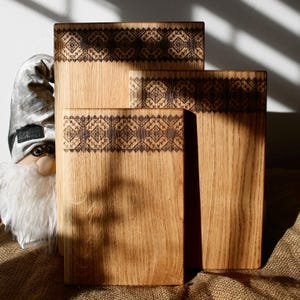 May include: Three wooden cutting boards of varying sizes. Each board is rectangular with a decorative, dark brown, geometric pattern etched along the top edge. The wood grain is visible, and the boards are arranged in a staggered display.