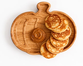 May include: A light brown wooden serving tray shaped like a pumpkin with a stem at the top. The tray holds a stack of six small, round, golden-brown pancakes and a single dollop of smooth, light brown caramel sauce placed on the wooden surface.