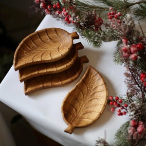 May include: Four wooden leaf-shaped serving dishes stacked on a white surface. The dishes are made of light brown wood and have a natural wood grain pattern.