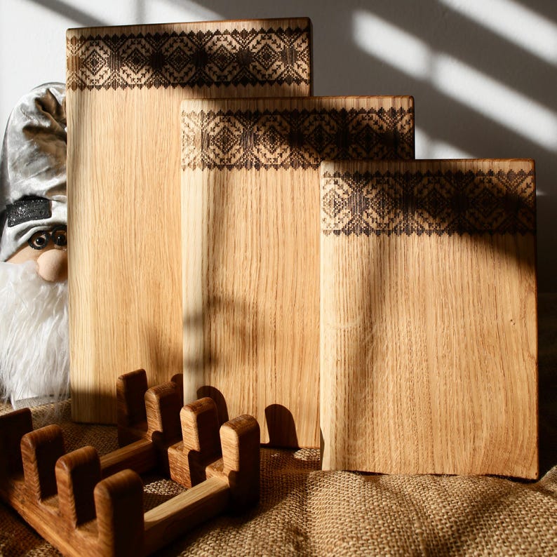 K&ouml;nnte beinhalten: Drei Holzschneidebretter mit einem dekorativen eingebrannten Design. Die Bretter sind &uuml;bereinander gestapelt und liegen auf einer Jutestoffoberfl&auml;che.