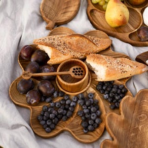 May include: Wooden serving platters with various compartments, filled with fresh fruit, bread, and a small wooden honey dipper. The arrangement includes plums, blueberries, grapes, and pears, perfect for a picnic or gathering.