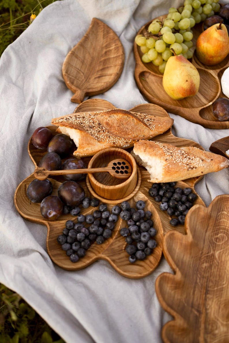 May include: Wooden serving platters with various compartments, filled with fresh fruit, bread, and a small wooden honey dipper. The arrangement includes plums, blueberries, grapes, and pears, perfect for a picnic or gathering.