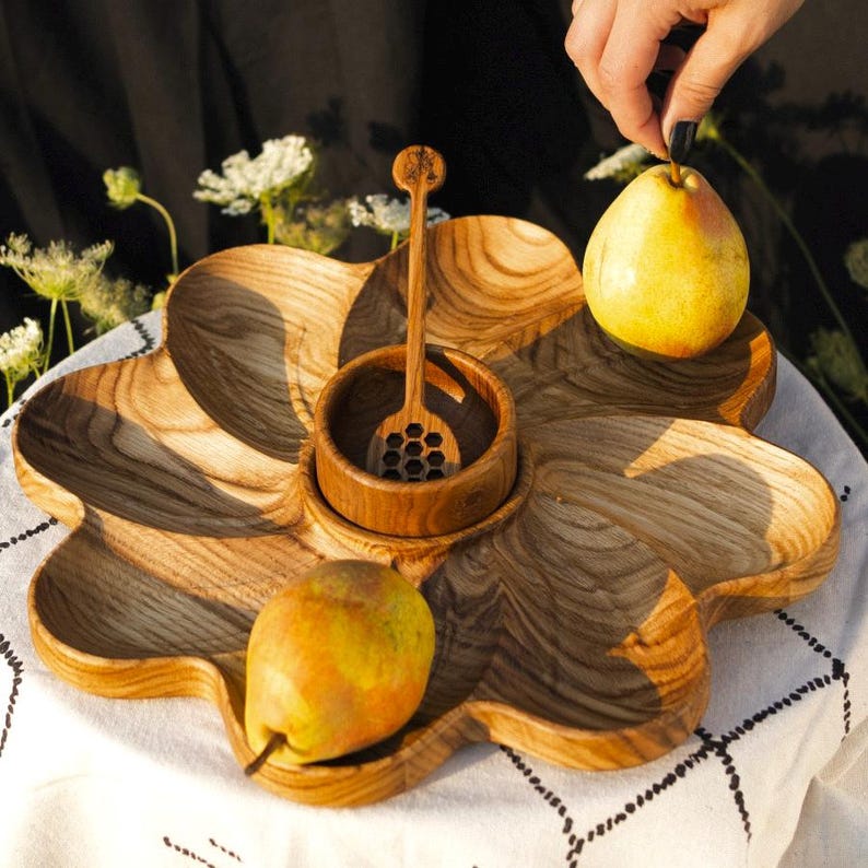 May include: A wooden serving tray with a flower-like design, featuring compartments for food. It includes a small bowl with a honey dipper and two pears. The tray is made of light brown wood and is set on a white tablecloth.