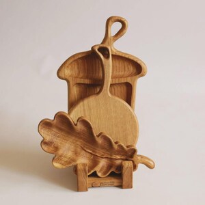 Può includere: Un set di tre taglieri e piatti da portata in legno marrone chiaro. Un tagliere ha la forma di una ghianda con manico, uno è un tagliere rotondo con manico e il terzo è un vassoio a forma di foglia di quercia con venature del legno visibili.