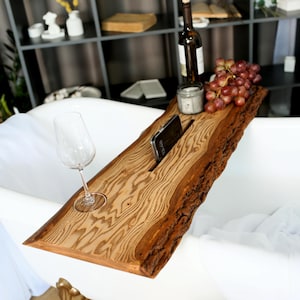 May include: A wooden bathtub tray with a live edge design. The tray has a slot for a phone, a wine glass holder, and is holding a wine bottle, grapes, and a candle. The wood grain is visible, and the tray is resting on a white bathtub.