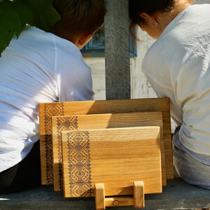 May include: Three wooden cutting boards of varying sizes, featuring a decorative geometric pattern on one side. The boards are displayed on a wooden stand, showcasing their natural wood grain and warm tones. The boards are made of wood.