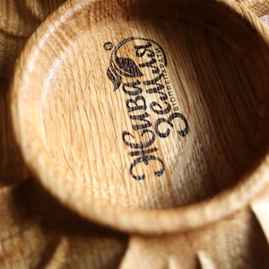 May include: Close-up of a wooden serving dish with a circular bowl and carved leaf-like details. The bowl features a logo with text in a foreign language. The wood grain is visible, and the dish has a natural, rustic appearance.