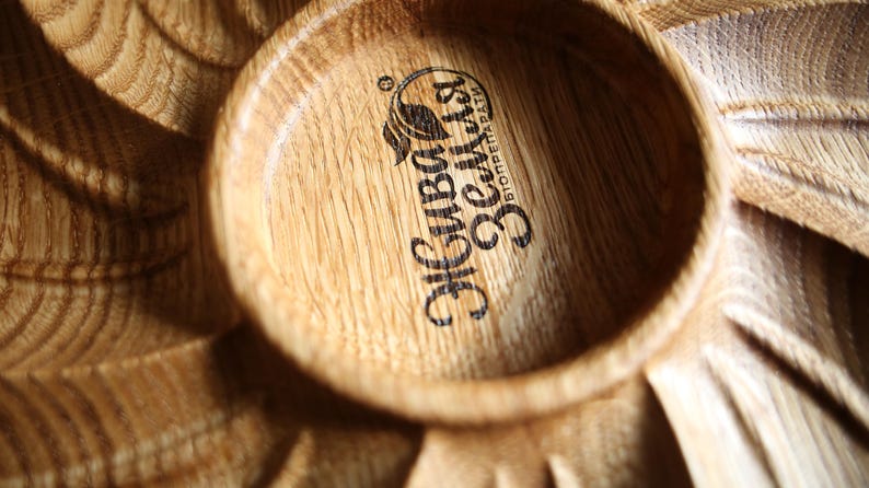 May include: Close-up of a wooden serving dish with a circular bowl and carved leaf-like details. The bowl features a logo with text in a foreign language. The wood grain is visible, and the dish has a natural, rustic appearance.