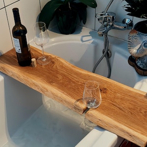 May include: A natural wood bathtub tray with a live edge, holding a wine bottle, two wine glasses, and a cork. The tray is made of light brown wood and spans the width of a white bathtub.