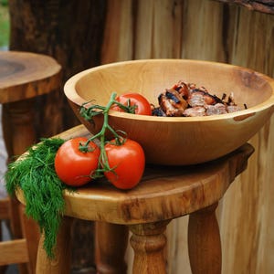 K&ouml;nnte beinhalten: Eine gro&szlig;e, runde Schale aus hellem Holz mit gegrillten Fleischst&uuml;cken, platziert auf einem Holzhocker neben einer Rispe aus drei leuchtend roten Tomaten und einem frischen Bund gr&uuml;ner Dill, die die nat&uuml;rliche Holzmaserung zeigt.