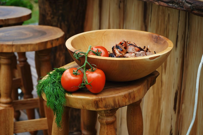 K&ouml;nnte beinhalten: Eine gro&szlig;e, runde Schale aus hellem Holz mit gegrillten Fleischst&uuml;cken, platziert auf einem Holzhocker neben einer Rispe aus drei leuchtend roten Tomaten und einem frischen Bund gr&uuml;ner Dill, die die nat&uuml;rliche Holzmaserung zeigt.