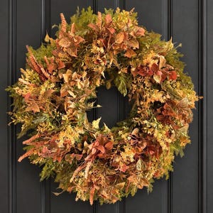 May include: A circular wreath composed of artificial foliage in shades of rust, orange, yellow, and deep green. The arrangement features a dense mix of textured leaves, small delicate flowers, and slender sprigs, creating a full, layered appearance.