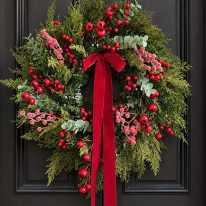 Op de afbeelding: Een ronde krans bestaande uit groen cederblad, afgewisseld met felrode bessen en lichtgroene eucalyptusbladeren. Een lang, felrood fluwelen lint is bovenaan tot een strik gebonden, met twee lange linten die in het midden naar beneden hangen.