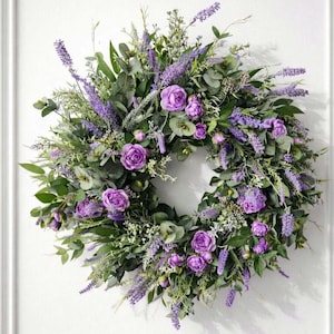 Könnte beinhalten: Ein runder Kranz mit violetten Rosen und Lavendelzweigen, durchsetzt mit verschiedenen grünen Eukalyptusblättern und Blattwerk. Das Arrangement ist dicht und geschichtet, was eine volle, abgerundete Form mit einer Mischung aus hellen und dunklen Grüntönen ergibt.