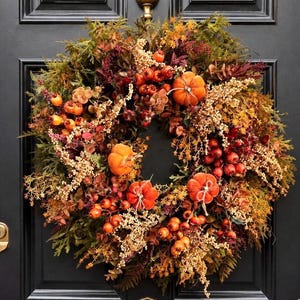 May include: A circular autumn wreath featuring orange pumpkins, clusters of red berries, dried foliage, and golden-brown leaves. The arrangement is dense with various textures of greenery and seasonal botanical elements, creating a full, rounded display.