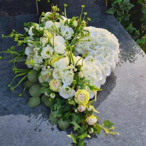 Könnte beinhalten: Ein herzförmiges Blumenarrangement aus zahlreichen weißen Rosen, kleinen hellgrünen Knospen und herabhängenden Efeublättern. Der Strauß besteht aus einer dichten Ansammlung weißer Blüten, die durch zartes grünes Blattwerk akzentuiert werden.