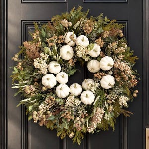 Peut inclure: Une couronne circulaire composée de nombreuses petites citrouilles blanches nichées parmi un arrangement dense de feuillage vert, d'herbes brunes séchées et de grappes de petites baies crème. L'objet décoratif est fixé sur une porte sombre.