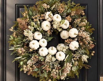 May include: A circular wreath featuring numerous small white pumpkins nestled among a dense arrangement of green foliage, dried brown grasses, and clusters of small cream-colored berries. The decorative piece is displayed on a dark door.