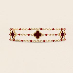 May include: A gold-colored bracelet featuring three red clover-shaped charms and red bead accents. The bracelet has a delicate chain design and is displayed against a neutral background.
