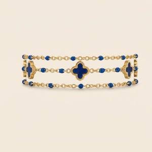 May include: A gold-toned bracelet featuring three rows of linked chains. Each row is adorned with small, round, blue beads. The bracelet has four-leaf clover-shaped charms with blue centers. The bracelet is displayed against a neutral background.