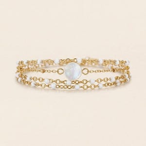May include: A gold-toned bracelet with three strands. The top and bottom strands feature small white beads, while the middle strand has a large, round, iridescent white stone. The bracelet is designed with delicate chain links.
