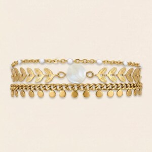 May include: Three-layer gold-toned bracelet set. The top layer features a delicate chain with white beads. The middle layer has leaf-shaped charms and a mother-of-pearl pendant. The bottom layer has a chain with small, dangling circular charms.