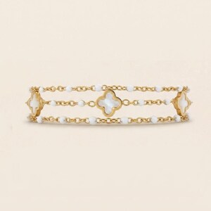May include: Gold-toned bracelet featuring three strands. Each strand has white beads and is connected by a gold chain. The bracelet has three clover-shaped charms with a white center. The bracelet is a jewelry item.