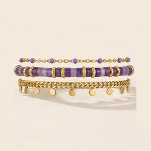 May include: Three stacked bracelets. The top bracelet is a delicate gold chain with small purple beads. The middle bracelet features purple and gold beads. The bottom bracelet is a gold chain with small, dangling gold discs.