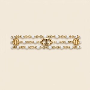 May include: Gold-toned bracelet featuring three strands. Each strand has white beads and a chain. The bracelet has three decorative gold pieces with a CD logo. The bracelet is displayed on a cream-colored background.