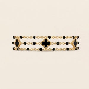 May include: A gold-colored bracelet featuring three rows of delicate chains. Each row is adorned with black beads and clover-shaped black accents. The bracelet has a symmetrical design and a polished finish.