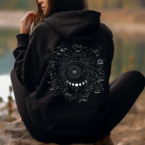 Puede incluir: Sudadera con capucha negra con un estampado blanco de un diseño celestial que presenta una fase lunar, estrellas y constelaciones. El diseño incluye los nombres de las constelaciones.