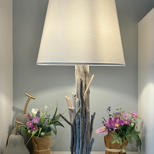 Op de afbeelding: Een decoratieve tafellamp met een drijfhouten voet en een witte lampenkap. De lamp heeft een zwarte cirkelvormige voet en wordt geflankeerd door bloemstukken in jute. Een uniek woondecoratie-item.