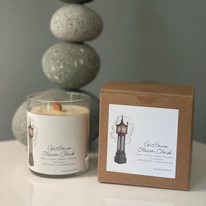 May include: A brown box with a white label featuring a drawing of a clock tower and the text "Gastown Steam Clock" with the scent description "Vanilla | Clove | Cream | Cinnamon". The box is next to a clear glass candle with a wooden wick and a white label with the text "Gastown Steam Clock" and the scent description "Vanilla | Clove | Cream | Cinnamon".