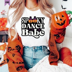 Könnte beinhalten: Ein weißes T-Shirt mit dem Text "Spooky Dance Babe" in schwarzen und orangen Buchstaben. Der Text ist mit zwei Kürbissen verziert, die Hexenhüte tragen.