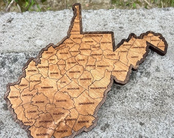 May include: A wooden map of West Virginia with each county outlined and labeled. The map is a dark brown color with a light brown wood grain.