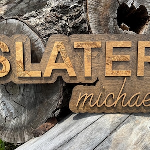May include: A layered wooden sign featuring the name SLATER in large block letters with engraved nature motifs including a deer, leaves, a bird, a fish, and a bear, with the name michael in a smaller cursive script layered below in wood.