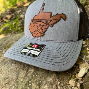 May include: A gray and black trucker hat with a brown leather patch of the state of West Virginia with a bridge over a river. The hat has the text "EST. 1970 RICHARDSON 112 TRUCKER ADJUSTABLE" on the label.