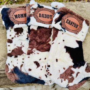 May include: Three Christmas stockings with a cowhide pattern in brown, black, and white. Each stocking has a brown leather patch with a name: "MOMM", "DADDY", and "EASTON". The stockings have a fluffy, faux fur texture.