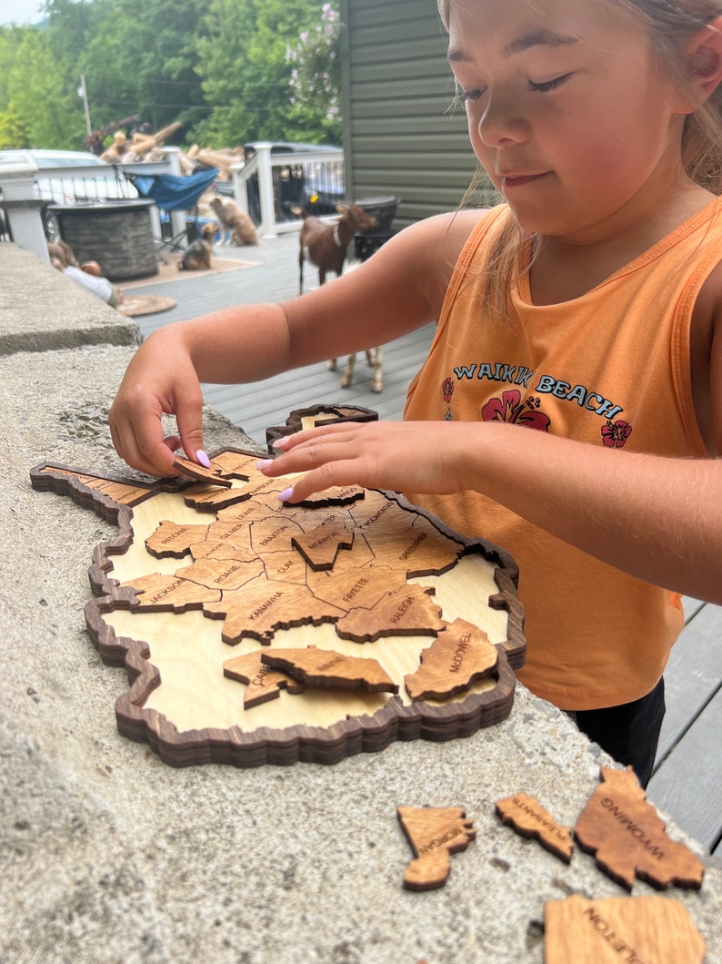 May include: A child is putting together a wooden puzzle of the state of West Virginia. The puzzle pieces are labelled with the names of the counties. The puzzle is on a grey concrete surface.