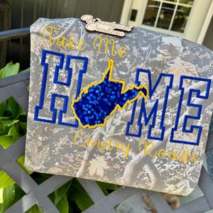 May include: A camouflage print t-shirt featuring the embroidered text Take Me HOME Country Roads in yellow and blue. The letter O in HOME is replaced by a blue sequined map of West Virginia outlined in yellow. A wooden logo tag is attached.