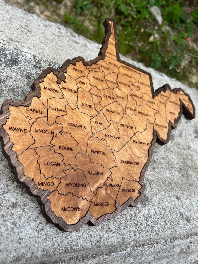 May include: A wooden map of West Virginia with the names of each county labelled. The map is made of light brown wood with dark brown borders around each county.