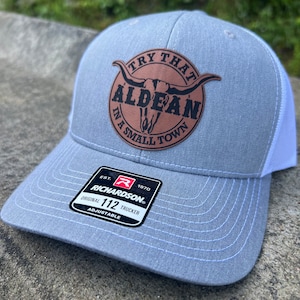 May include: A gray and white trucker hat with a brown leather patch that says "Try That Aldean In A Small Town". The patch features a black silhouette of a bull's head with horns. The hat is adjustable and has a Richardson 112 label.