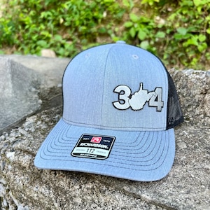 May include: A gray and black trucker hat with a white embroidered patch featuring the number 304 and the outline of the state of West Virginia. The hat is adjustable and has a black mesh back.
