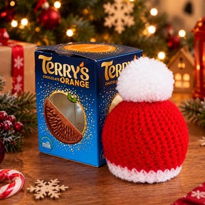 Könnte beinhalten: Eine blaue Pappschachtel von Terry's Chocolate Orange mit einem kreisförmigen Ausschnitt, der ein Schokoladensegment zeigt, neben einer kleinen, runden, roten Häkelmütze mit weißem Rand und einem großen weißen Bommel.